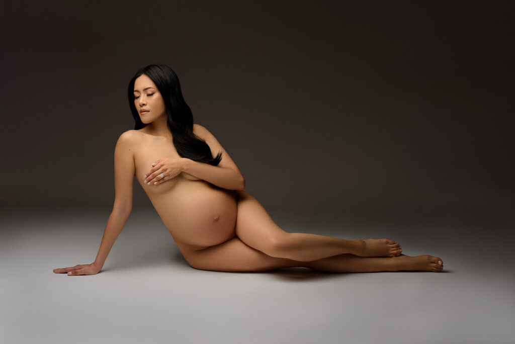 Moody maternity portrait of a woman posing nude in front of a dark grey backdrop