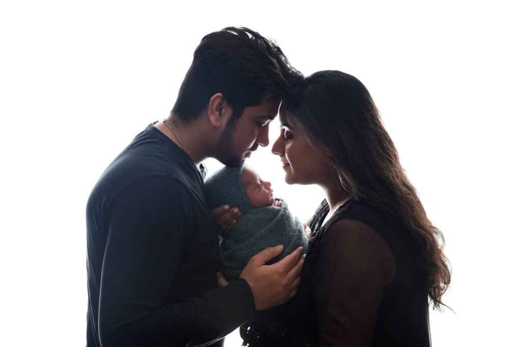 A couple faces each other, gently touching foreheads while lovingly holding a newborn baby swaddled in a blue blanket. The background is a bright white, emphasizing the intimate and heartwarming moment captured in this beautiful piece of newborn photography. Image by NYC Maternity, Newborn & Baby Photographer, Brilianna Photography.