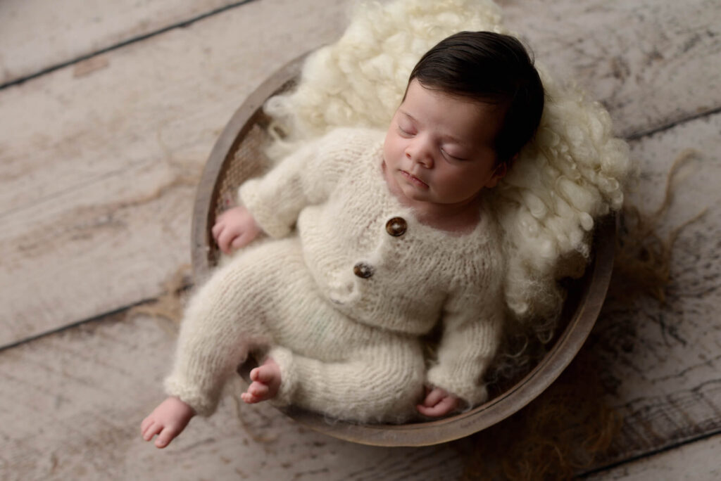 Portrait of a newborn baby boy resting peacefully in a white sweater, nestled within a bowl.