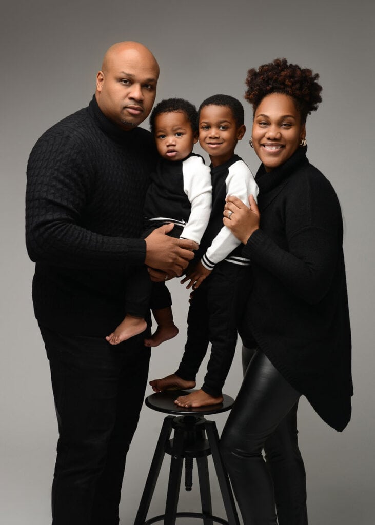 A family of four poses for a portrait against a gray background. The man on the left holds a baby, while the older child stands barefoot on a black stool between the parents. Both children are wearing matching black and white outfits. The woman on the right smiles warmly, capturing this moment in photography. Image by NYC Maternity, Newborn & Baby Photographer, Brilianna Photography.