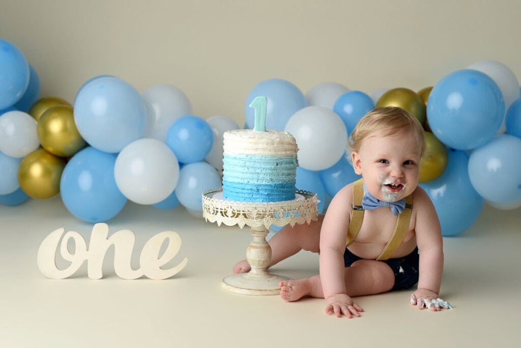 A baby dressed in a blue bow tie and shorts sits on the floor beside a small cake with a "1" topper, perfect for infant pictures. The baby has cake on their face and fingers. Blue and gold balloons are in the background, and a wooden "one" sign is placed to the left, making it an adorable scene of baby photography. Image by NYC Maternity, Newborn & Baby Photographer, Brilianna Photography.