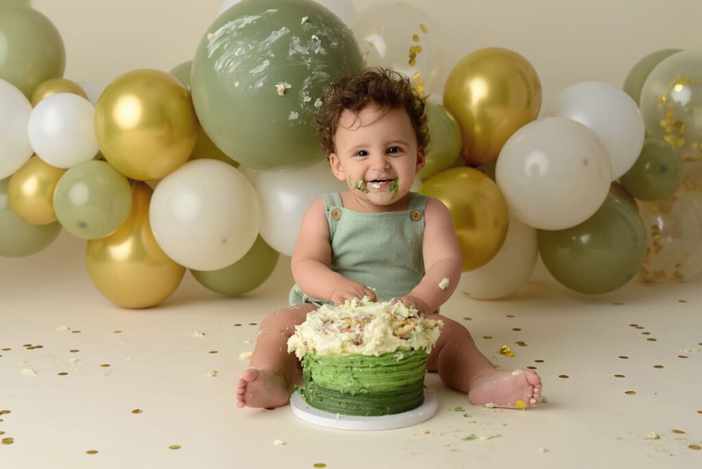 A baby sits on the floor, happily covered in cake, surrounded by green, gold, and white balloons. The baby wears a light green outfit and enjoys a messy, green and white frosted cake. Confetti and cake crumbs are scattered on the floor—a perfect moment of baby photography. Image by NYC Maternity, Newborn & Baby Photographer, Brilianna Photography.