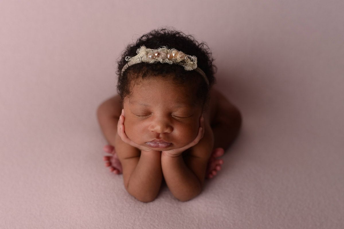 A newborn baby with curly hair sleeps peacefully, propped up on their hands. The baby wears a delicate flowered headband and is posed against a soft, pink background, creating a serene and tender scene—an ideal moment captured by the art of newborn photography. Image by NYC Maternity, Newborn & Baby Photographer, Brilianna Photography.