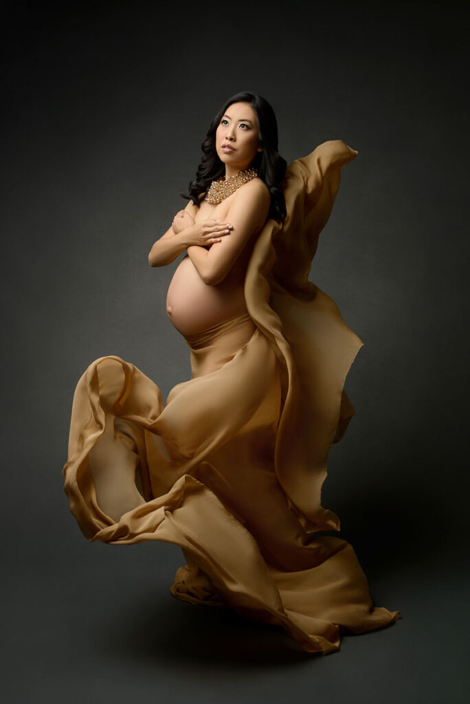 Darkly lit maternity portrait of pregnant woman in a gold gown draped loosely around her stomach