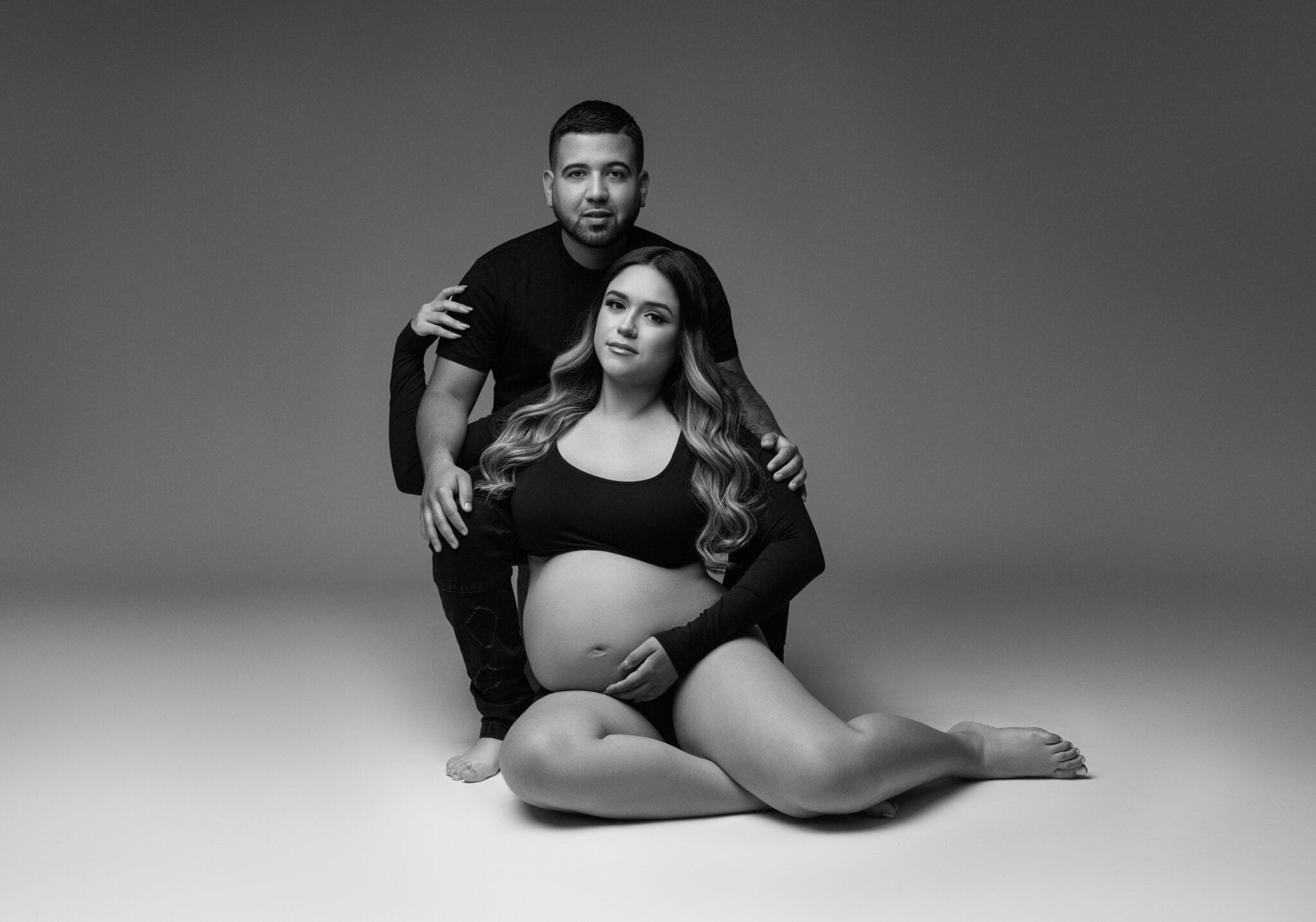A black-and-white photo of a pregnant woman sitting on the floor with legs crossed, cradling her belly, with a man kneeling behind her, hands on her shoulders. Both wear black outfits, creating an intimate portrait that guides viewers to the tender investment in their growing family against a plain studio backdrop. Image by NYC Maternity, Newborn & Baby Photographer, Brilianna Photography.