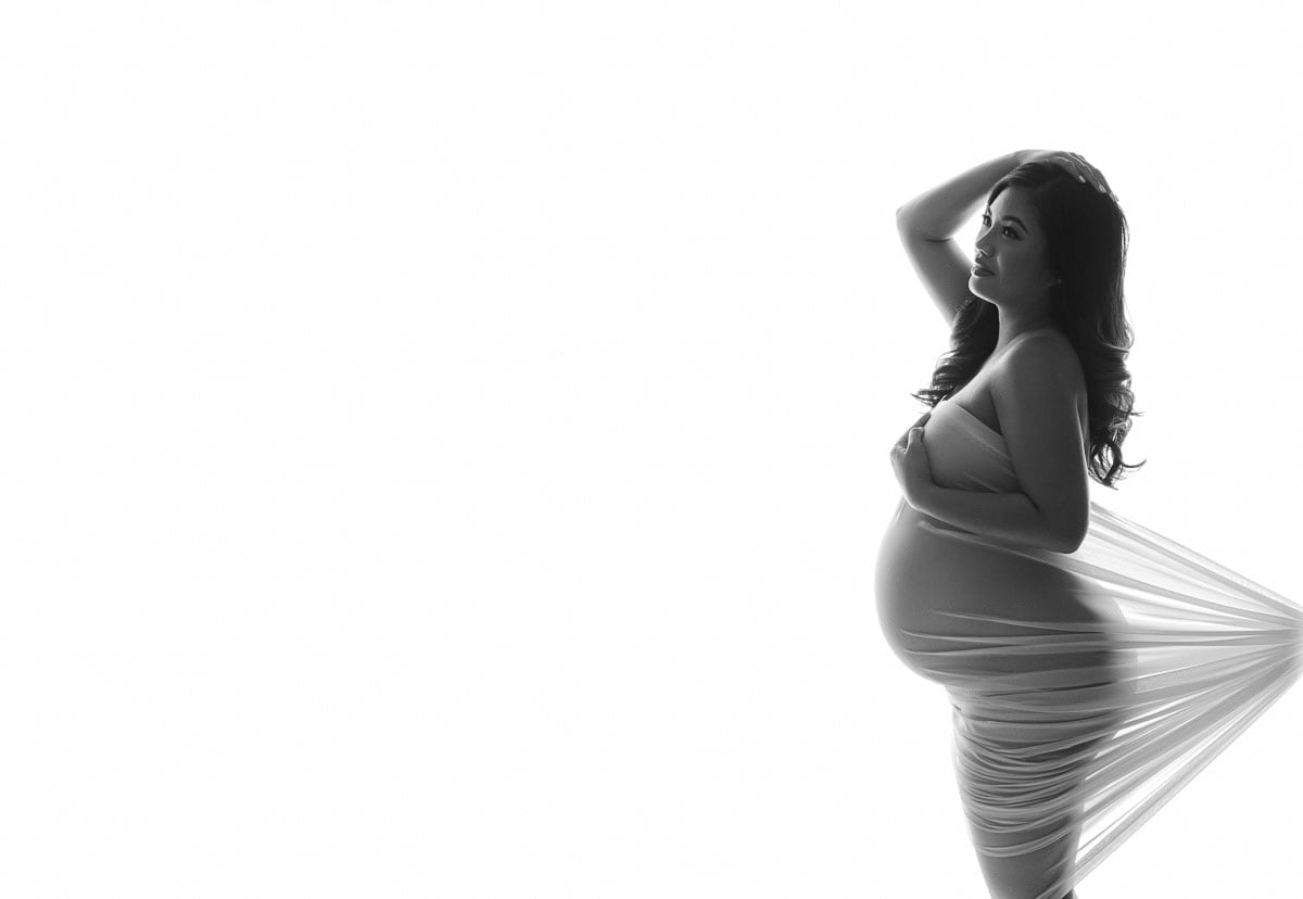 A pregnant woman is posing in a side profile, wrapped in a sheer fabric that accentuates her baby bump. She has one hand resting on her head and the other on her belly. The background is completely white, making the black-and-white image high in contrast. Image by NYC Maternity, Newborn & Baby Photographer, Brilianna Photography.