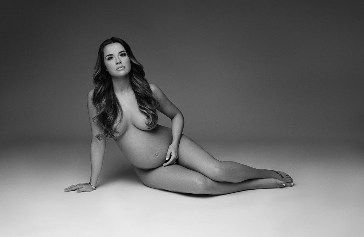 A black and white photo of a pregnant woman sitting on the floor. She is nude and posed gracefully with her body partially turned to the side, one hand resting on the floor and the other cradling her baby bump. Her long hair cascades over her shoulders. Image by NYC Maternity, Newborn & Baby Photographer, Brilianna Photography.