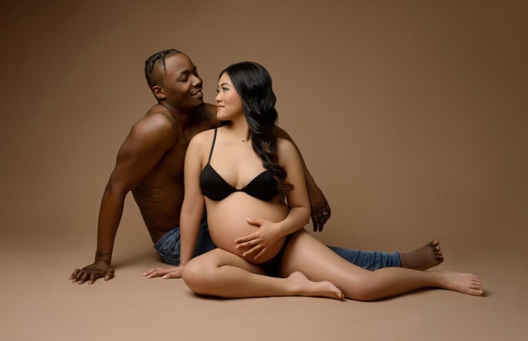 A pregnant woman in a black bra and underwear sits on the floor, cradling her belly with one hand. A shirtless man with jeans sits behind her, leaning back on his hands, smiling at her. Both are seated on a plain brown background. Image by NYC Maternity, Newborn & Baby Photographer, Brilianna Photography.