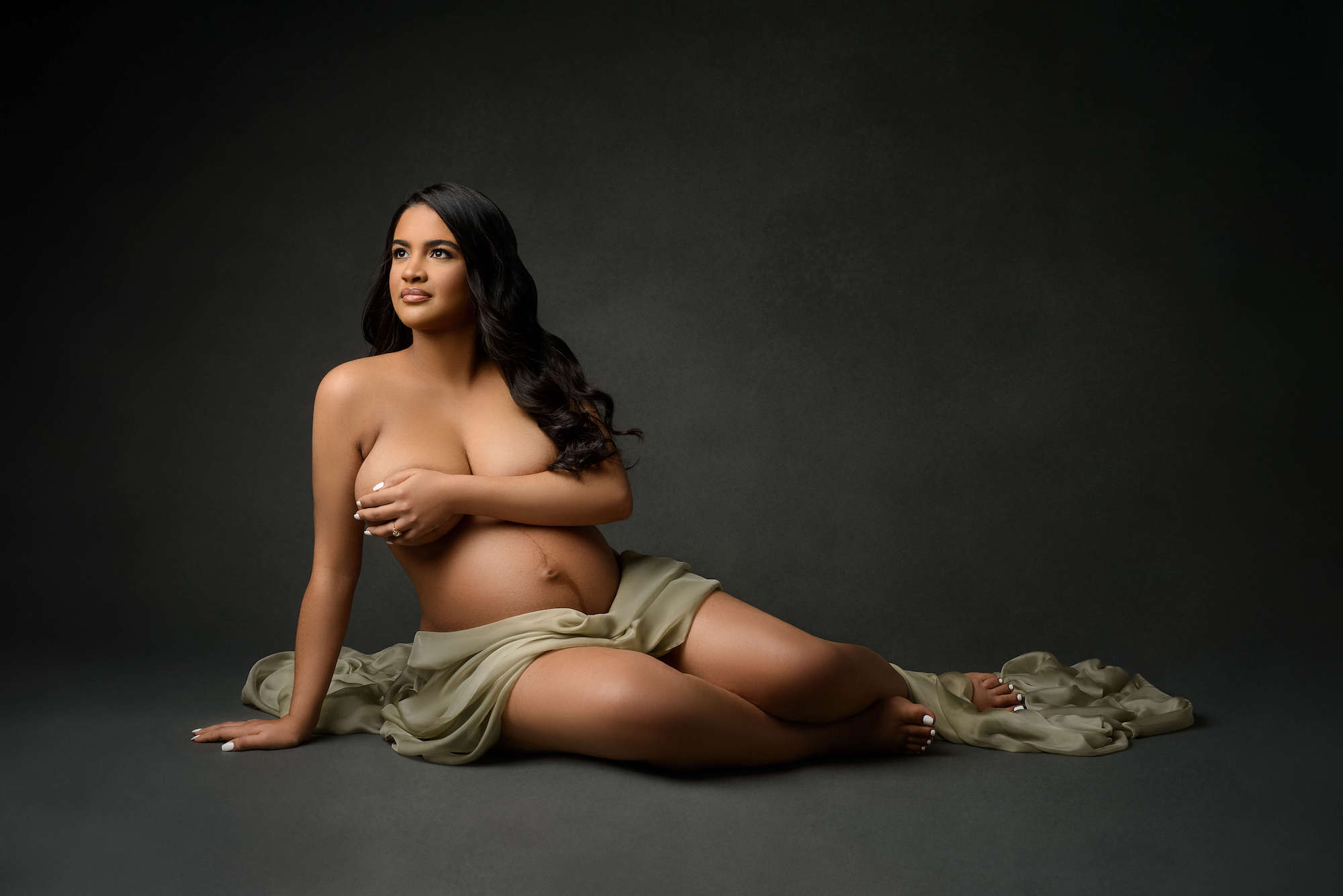 Editorial styled maternity portrait of woman sitting on floor draped in green cloth on a grey backdrop. 