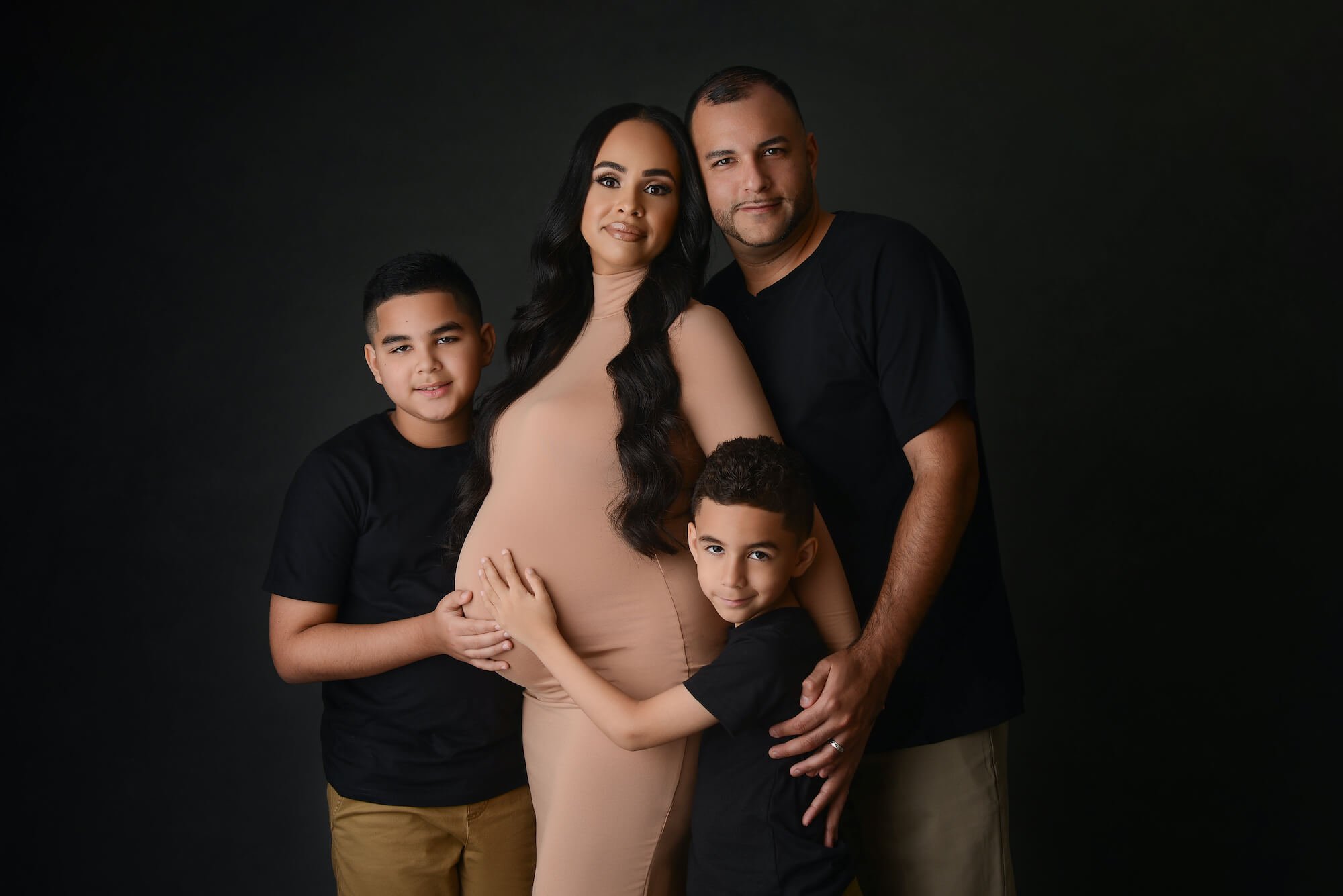 A pregnant woman in a beige dress poses with her family against a dark backdrop. Her husband stands beside her in a black shirt and khaki pants, while their two young sons, also in black shirts, stand in front—one hugging his mother and the other holding her hand. This beautiful maternity photography was captured in New York City. Image by NYC Maternity, Newborn & Baby Photographer, Brilianna Photography.