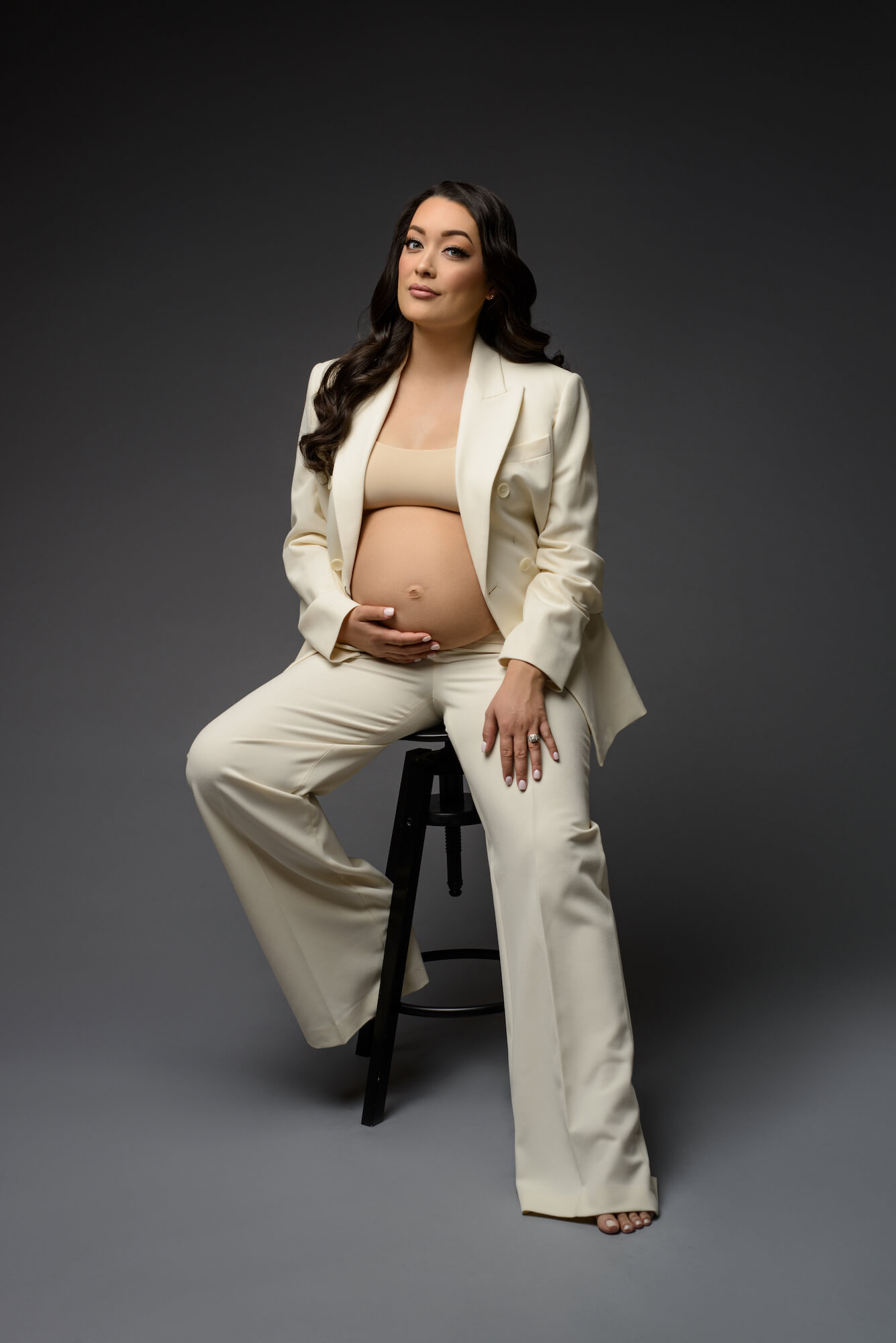 Classy maternity portrait of brunette sitting on stool wearing white business suit with the coat open to show off the baby bump. 