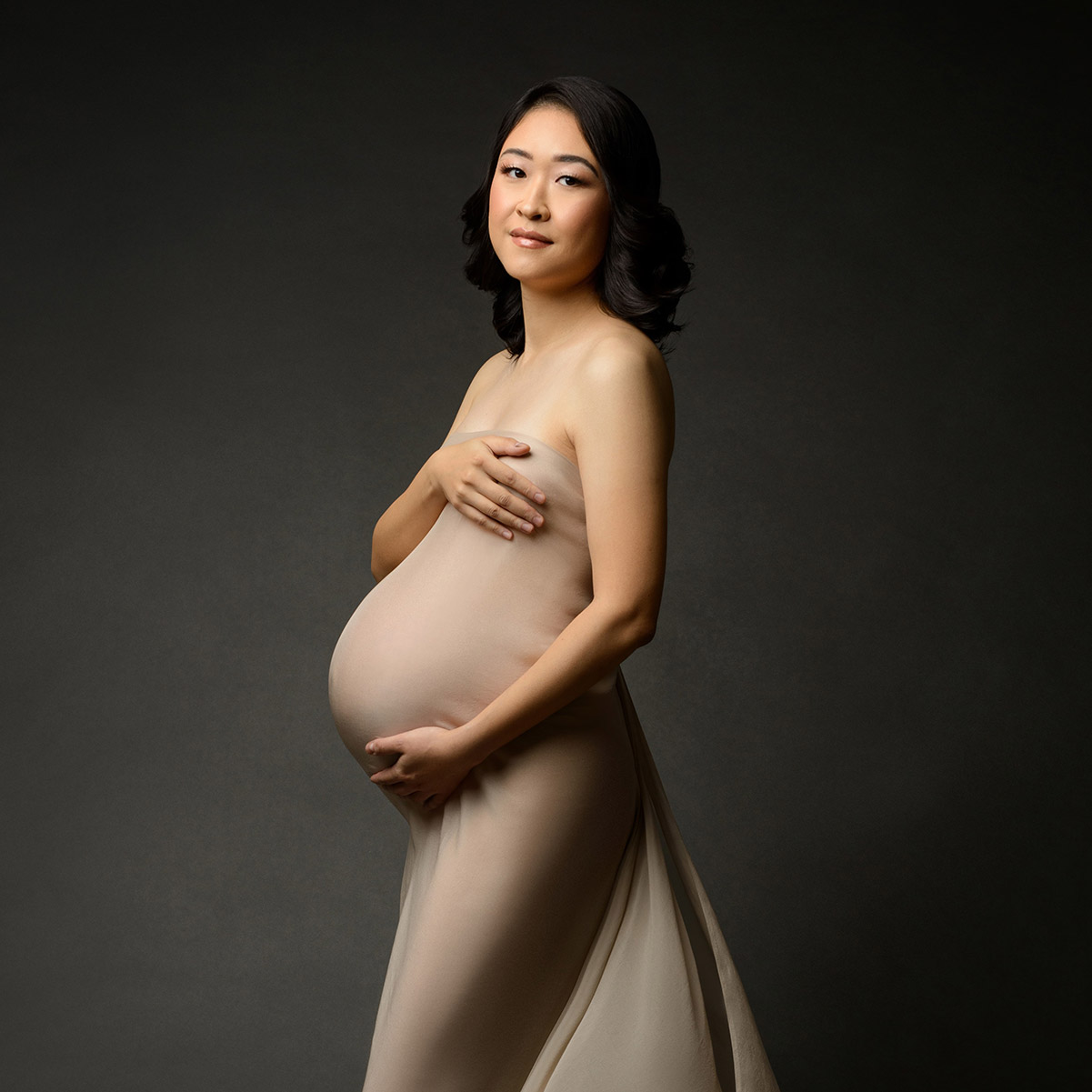 Maternity portrait of Asian woman, posing and smiling at the camera, wrapped tightly in a beige fabric.