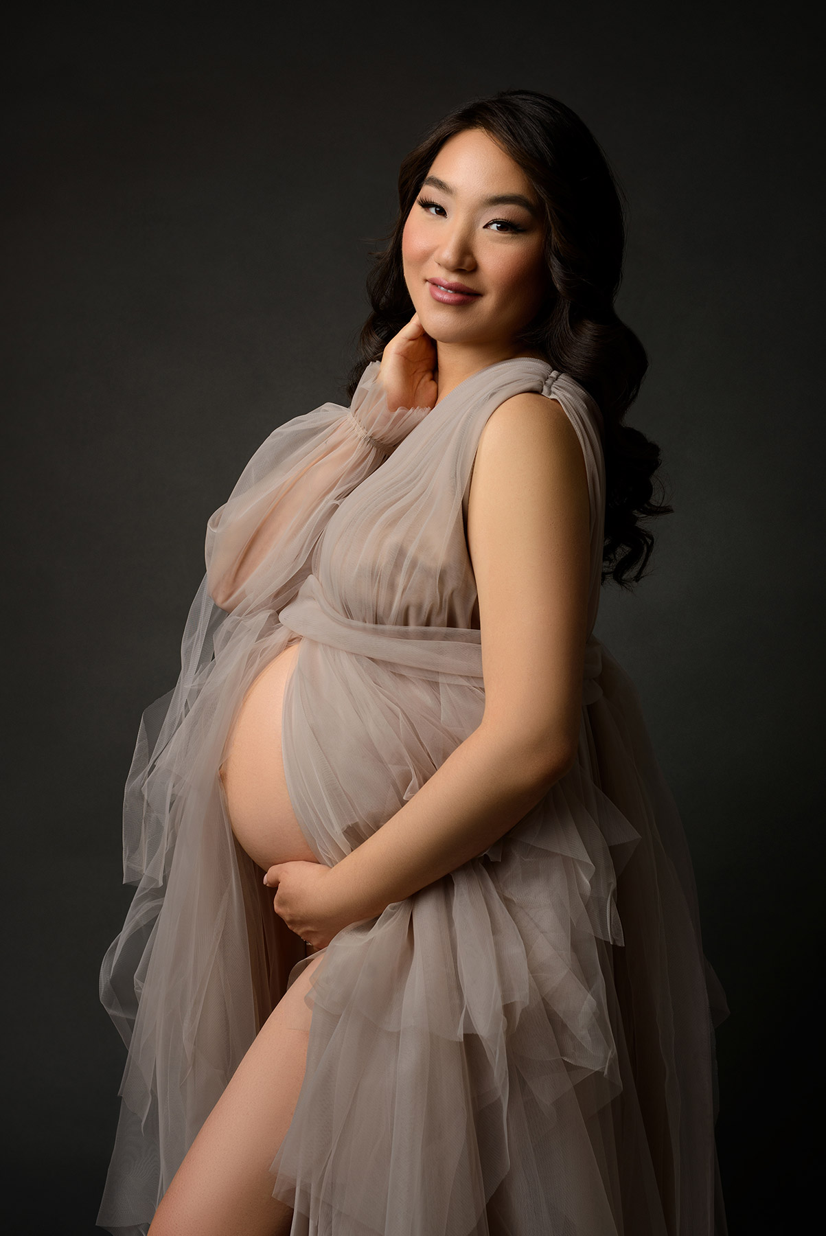 Luxurious maternity photo of Asian woman wearing tulle gown and smiling at the camera on a dark grey back drop.