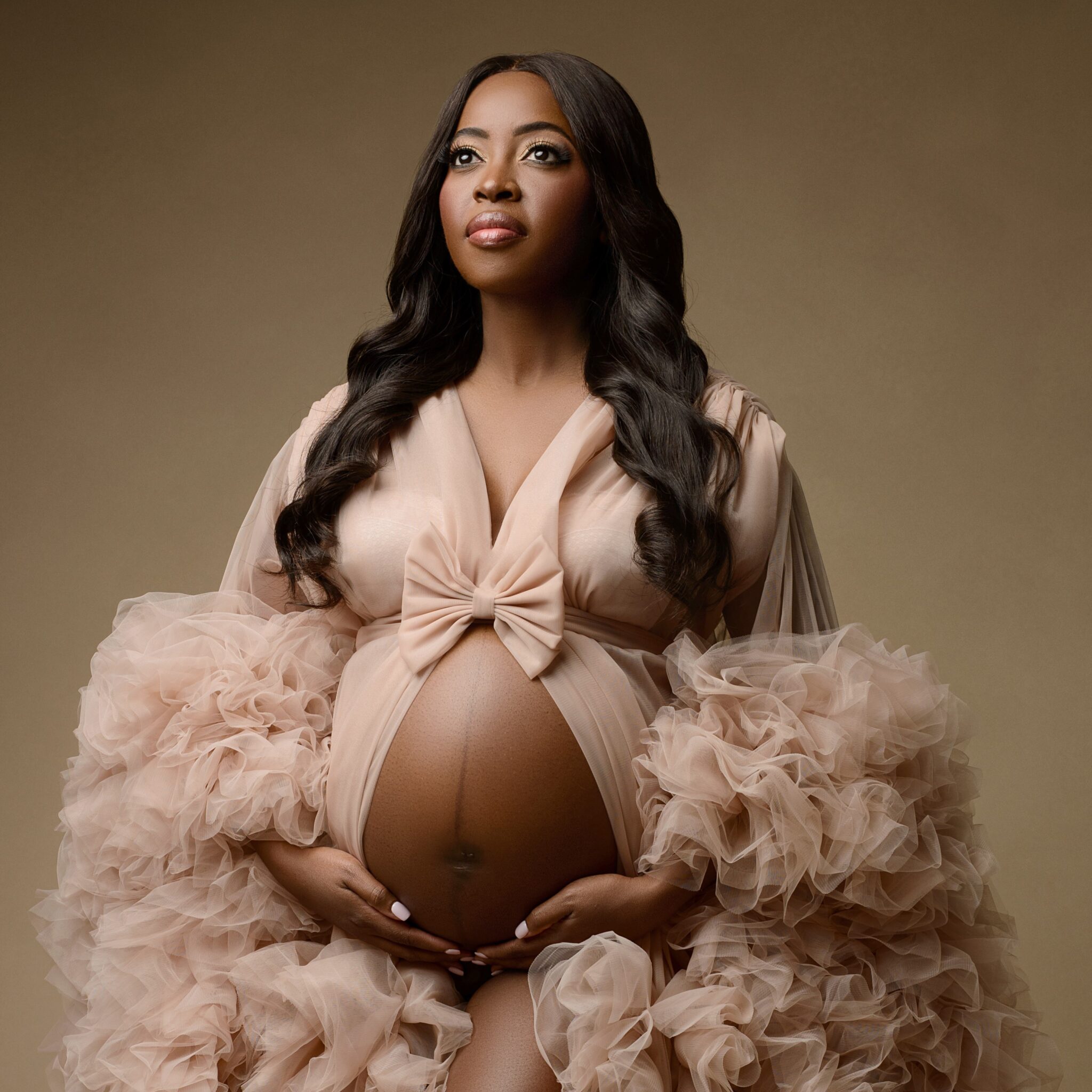Maternity portrait of black woman holding her baby bump poking thru a luxurious dusty-pink robe made of tulle with ruffles at the sleeves.