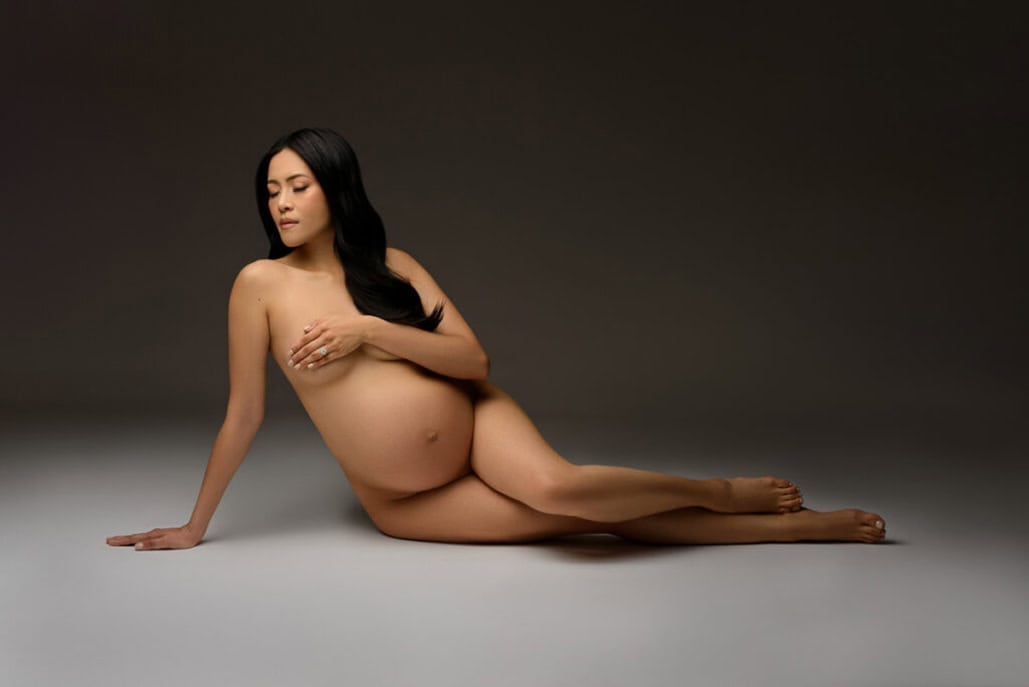 Moody maternity portrait of a woman posing nude in front of a dark grey backdrop