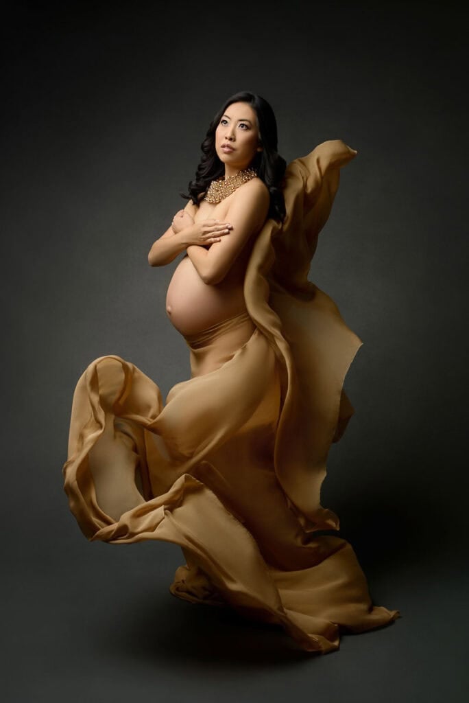 Darkly lit maternity portrait of pregnant woman in a gold gown draped loosely around her stomach