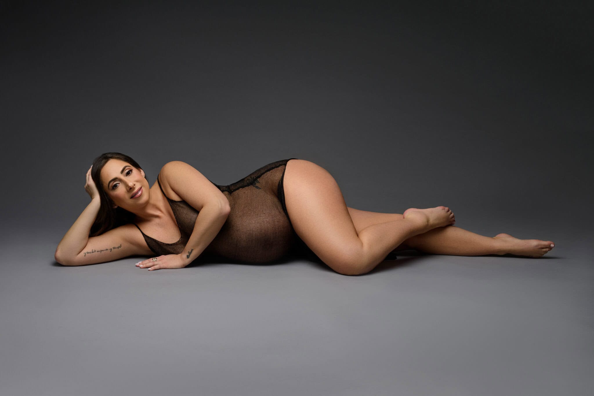 In a captivating piece of maternity photography, a pregnant woman in a sheer black bodysuit lies on her side against a gray surface with a dark gray background. Smiling and supporting her head with one hand, she displays a visible tattoo on her partially extended arm. Image by NYC Maternity, Newborn & Baby Photographer, Brilianna Photography.