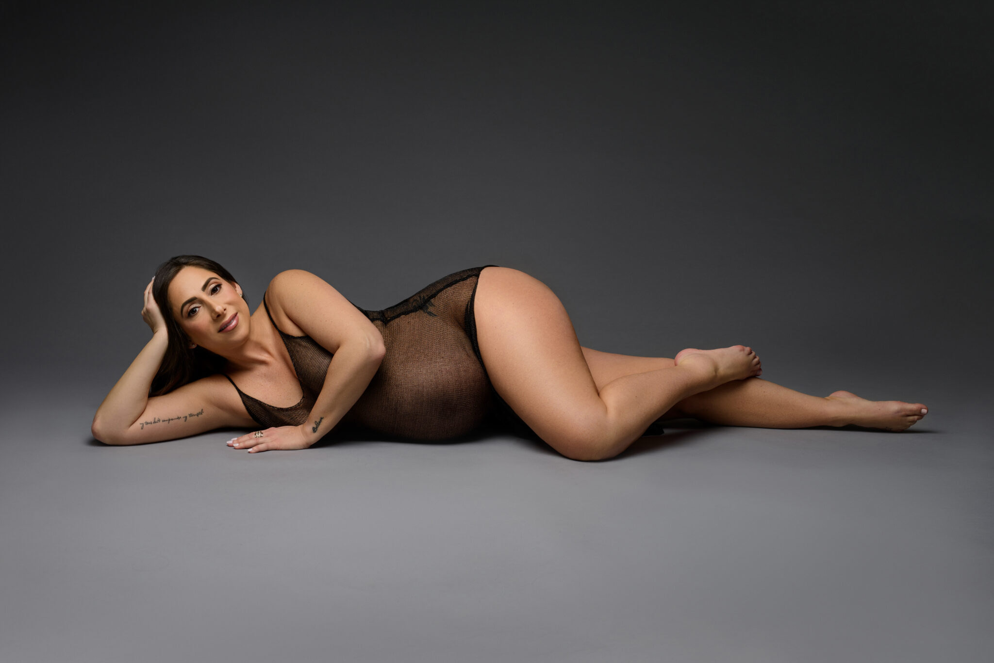 Portrait of a pregnant woman in a sleek sheer black body suit lying on the floor, exuding elegance and confidence.