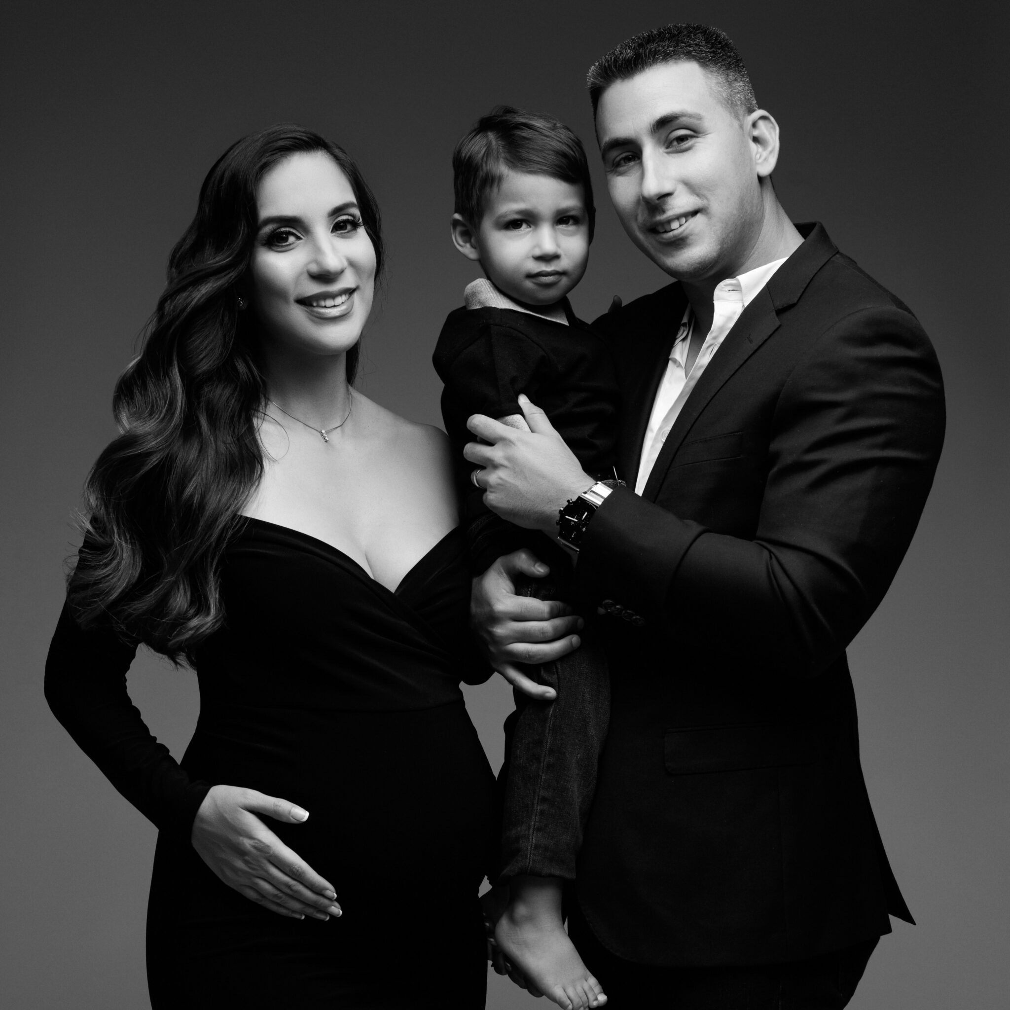 Black and white maternity portrait of family wearing all black formal attire, with the father holding the son and the expecting mother holding a hand on her baby bump.