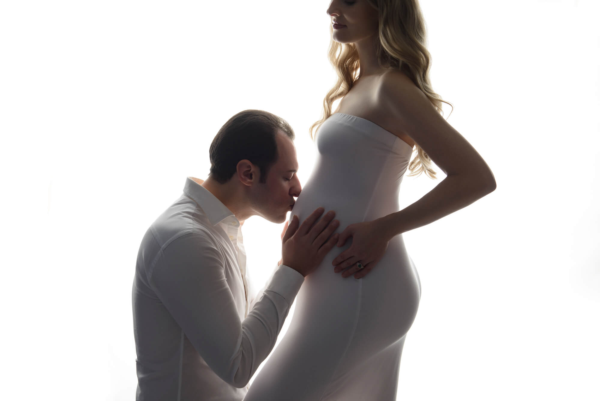 Maternity portrait on clean white background of couple. The man kneels down to kiss the woman's baby bump.