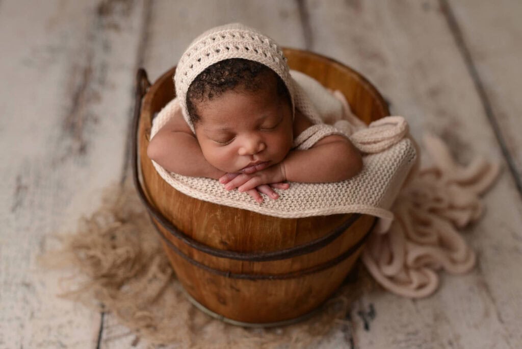 A newborn baby sleeps peacefully, head resting on folded arms, inside a wooden bucket. The baby is wrapped in a cream-colored knit blanket and wears a matching knit bonnet. Captured by a talented Manhattan Maternity Photographer, the bucket sits on a rustic wooden floor with textured cloth underneath. Image by NYC Maternity, Newborn & Baby Photographer, Brilianna Photography.