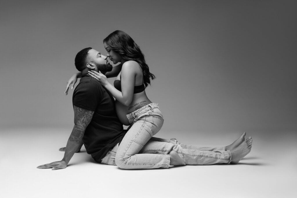 A black and white maternity photo captures a pregnant woman and a man sitting on the floor in New York City. The woman, wearing a crop top and unbuttoned jeans, sits on the man's lap, gently holding his face. The man, in a T-shirt and jeans, supports her with one hand on the floor as they share an intimate moment. Image by NYC Maternity, Newborn & Baby Photographer, Brilianna Photography.