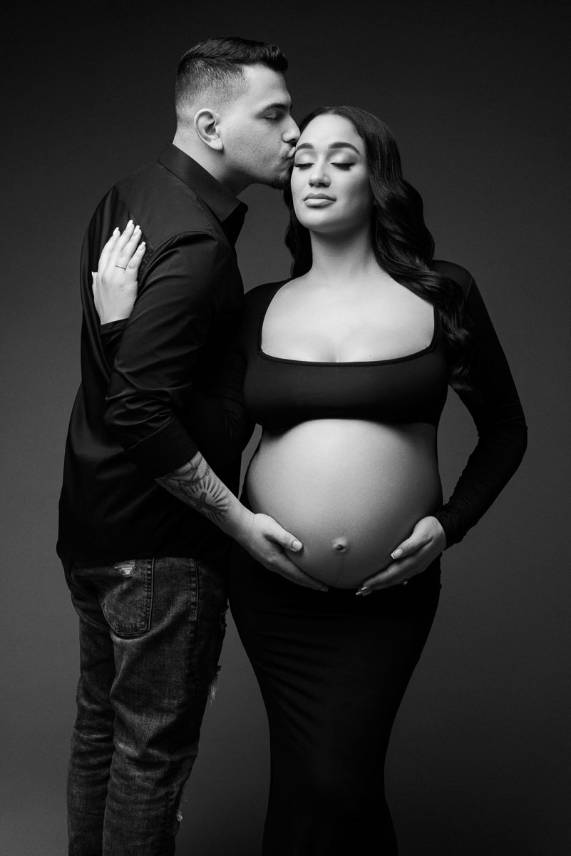 Professional maternity portrait of couple in black and white. The man gives the expecting mother a kiss on the forehead while holding her baby bump.