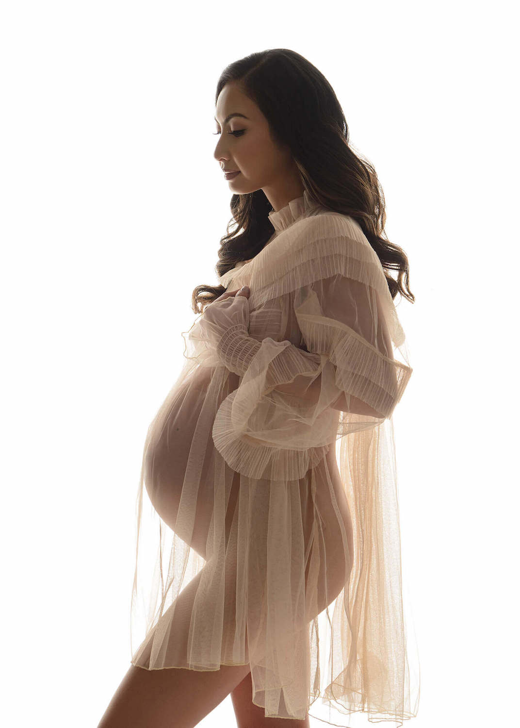 Luxurious maternity photo of brunette woman wearing tulle gown on a solid white backdrop.