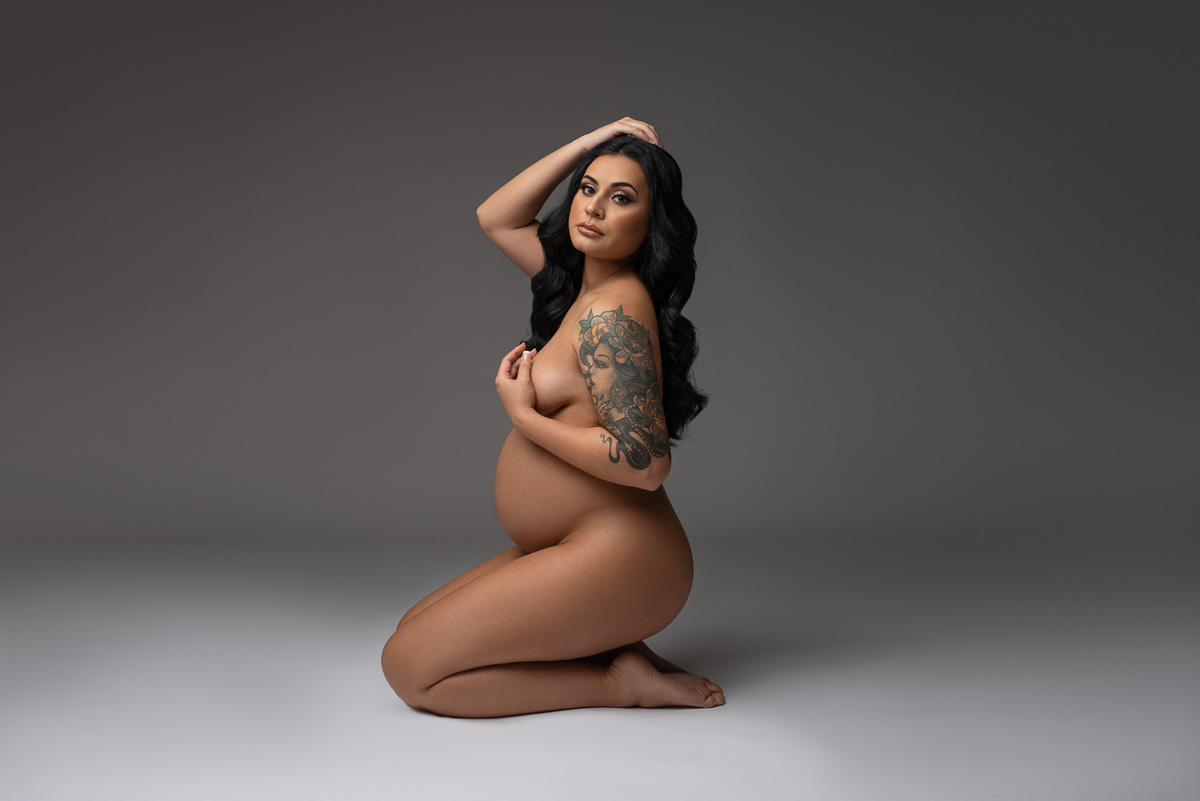 Classy implied nude maternity portrait, the expected mother is kneeling and facing the camera against a cool grey backdrop. 