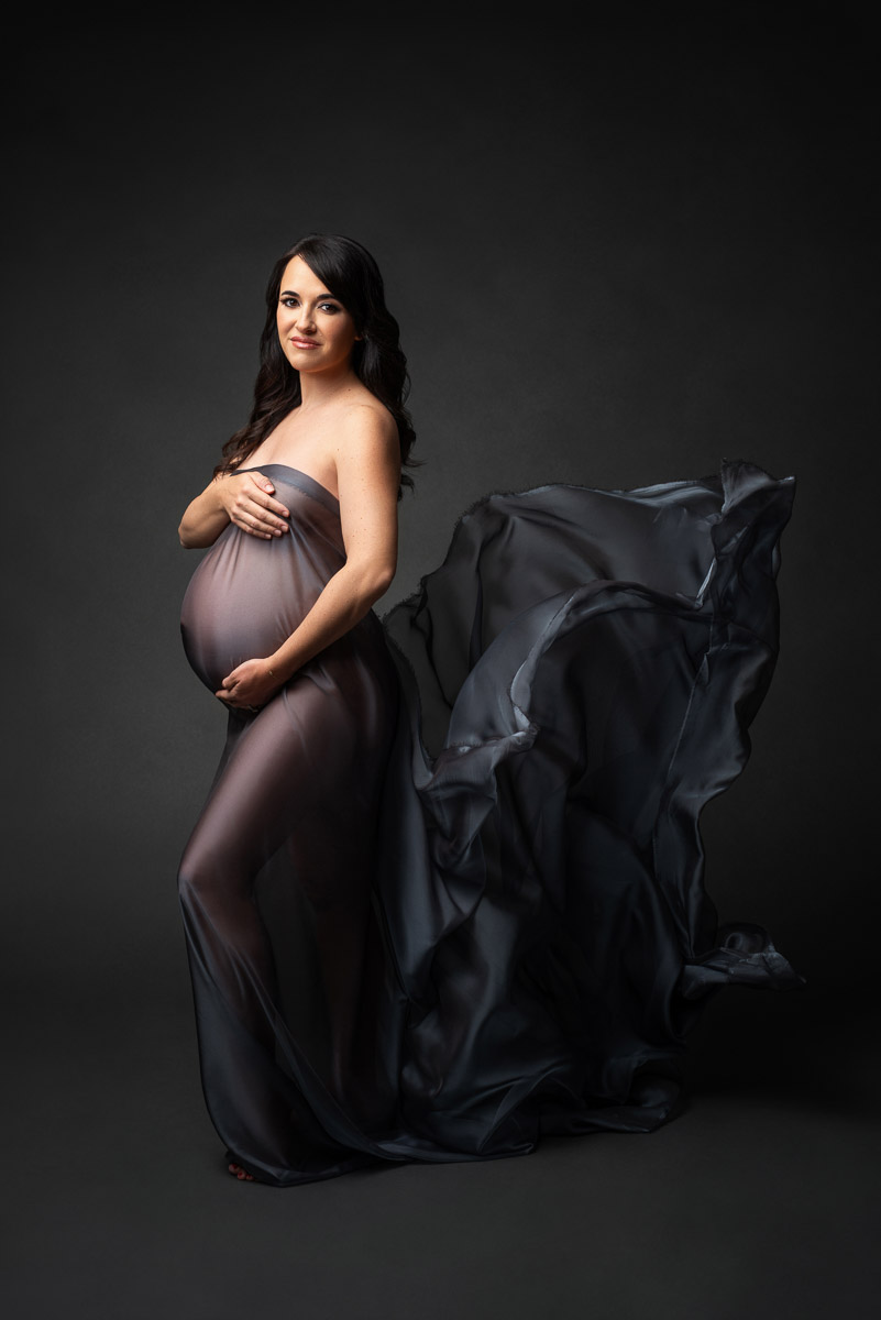 Stunning maternity portrait of woman smiling, standing, and wrapped in a sheer black cloth that is flowing behind her.